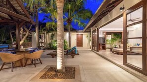 A covered outdoor area with a palm tree, wicker furniture, and a building with large windows. - Aqua Vista 28: 3BR Thai Style Villa (Koh Samui)