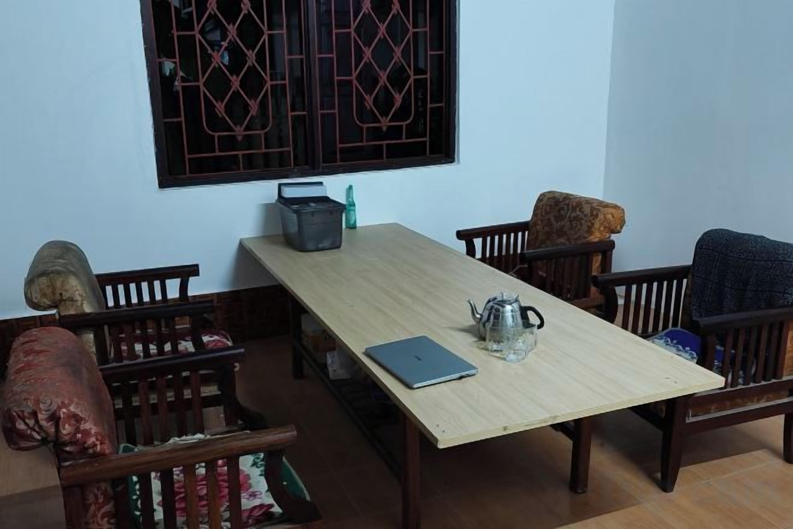 A dining area with a wooden table, chairs, a laptop, a teapot, and a window with a decorative grille.