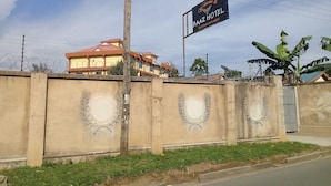 A Baaz Hotel sign with a welcoming message, a concrete wall with decorative leaf patterns, and a building with a red roof in the background. - Baaz Hotel Kakamega (Kakamega)
