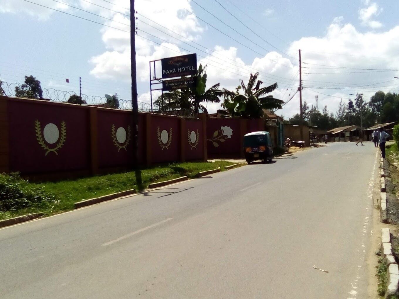 Une route avec un panneau indiquant l’hôtel Baaz, un mur rouge orné d’emblèmes circulaires et un tuk-tuk sur la route.