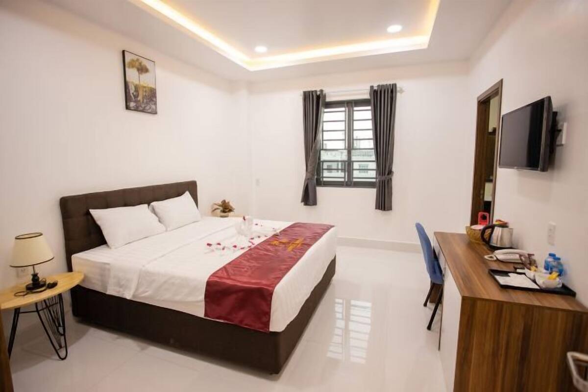 A hotel room with a bed, a desk, a chair, a TV, a window with curtains, and a painting on the wall.