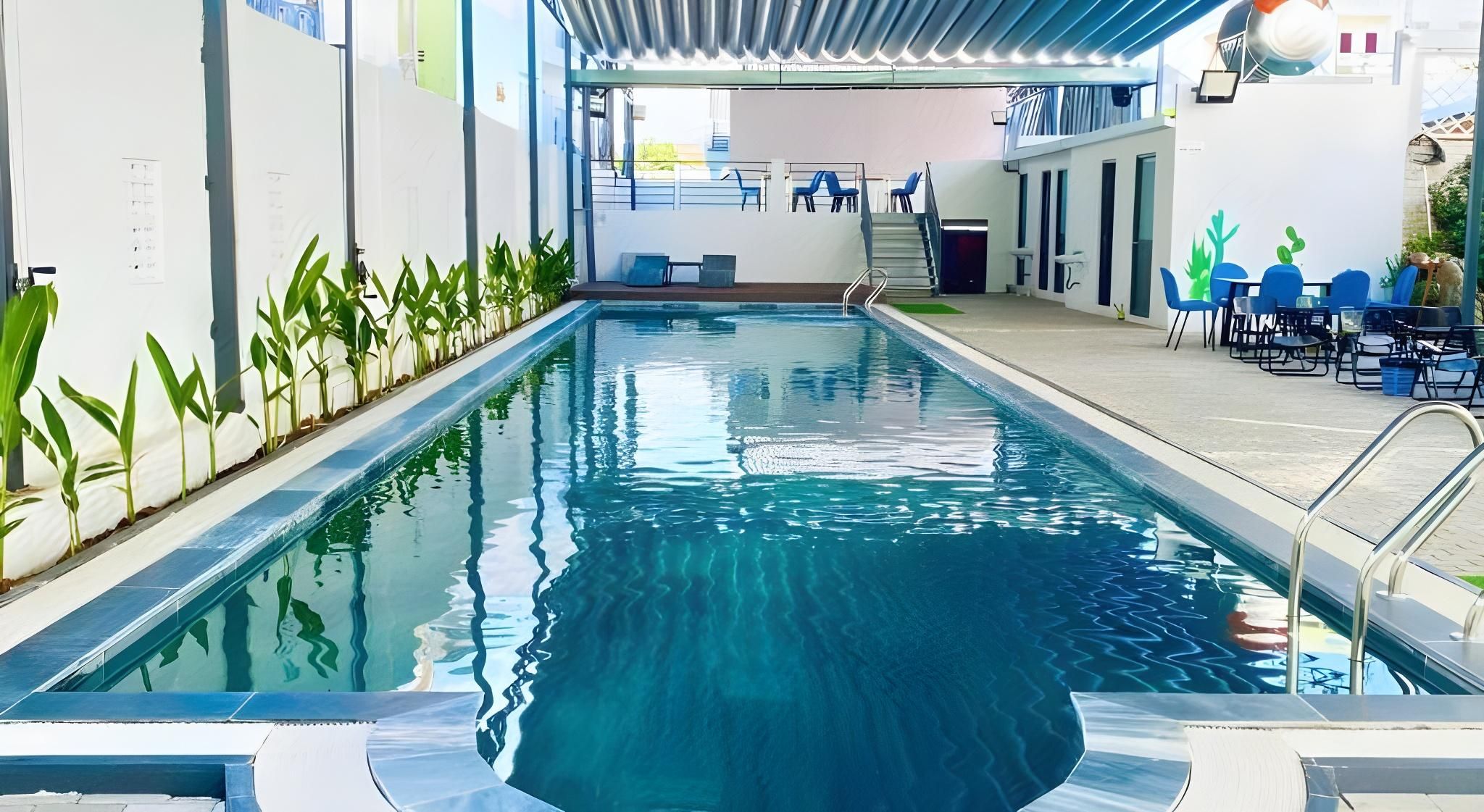 An indoor swimming pool with clear water, surrounded by walls with plants, and a seating area with chairs and tables.