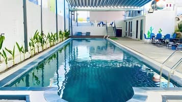 An indoor swimming pool with clear water, surrounded by walls with plants, and a seating area with chairs and tables.