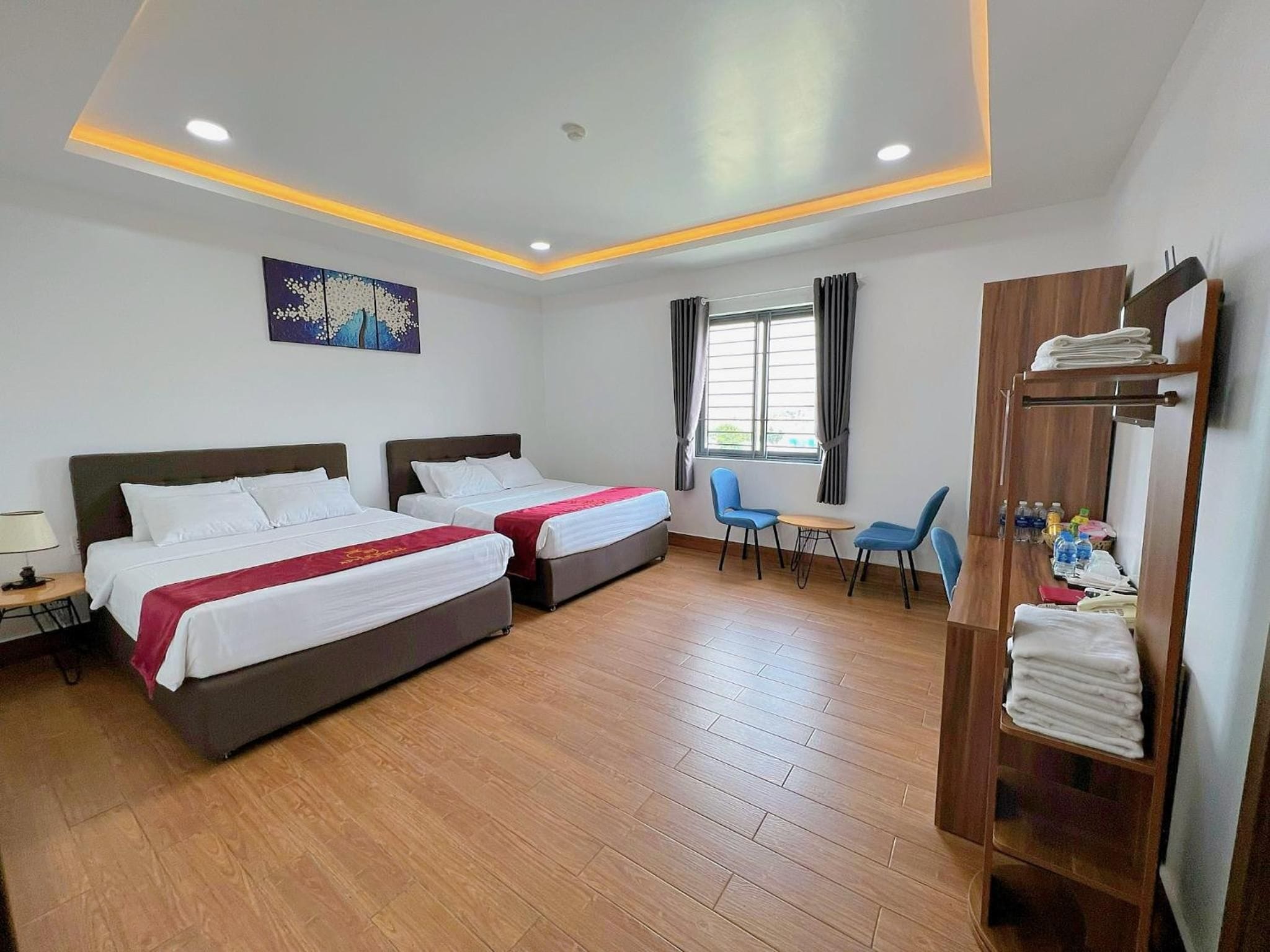 A hotel room with two beds, a wooden floor, a desk with a lamp, a chair, a window with curtains, and a wooden wardrobe with towels.