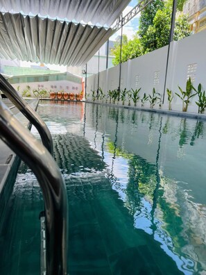 A swimming pool with clear water, surrounded by a white wall with small windows and plants. - Apple Hotel (Can Tho)
