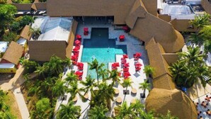 Vista aérea de un complejo turístico con una piscina central rodeada de sombrillas rojas y tumbonas, palmeras y estructuras con techo de paja.