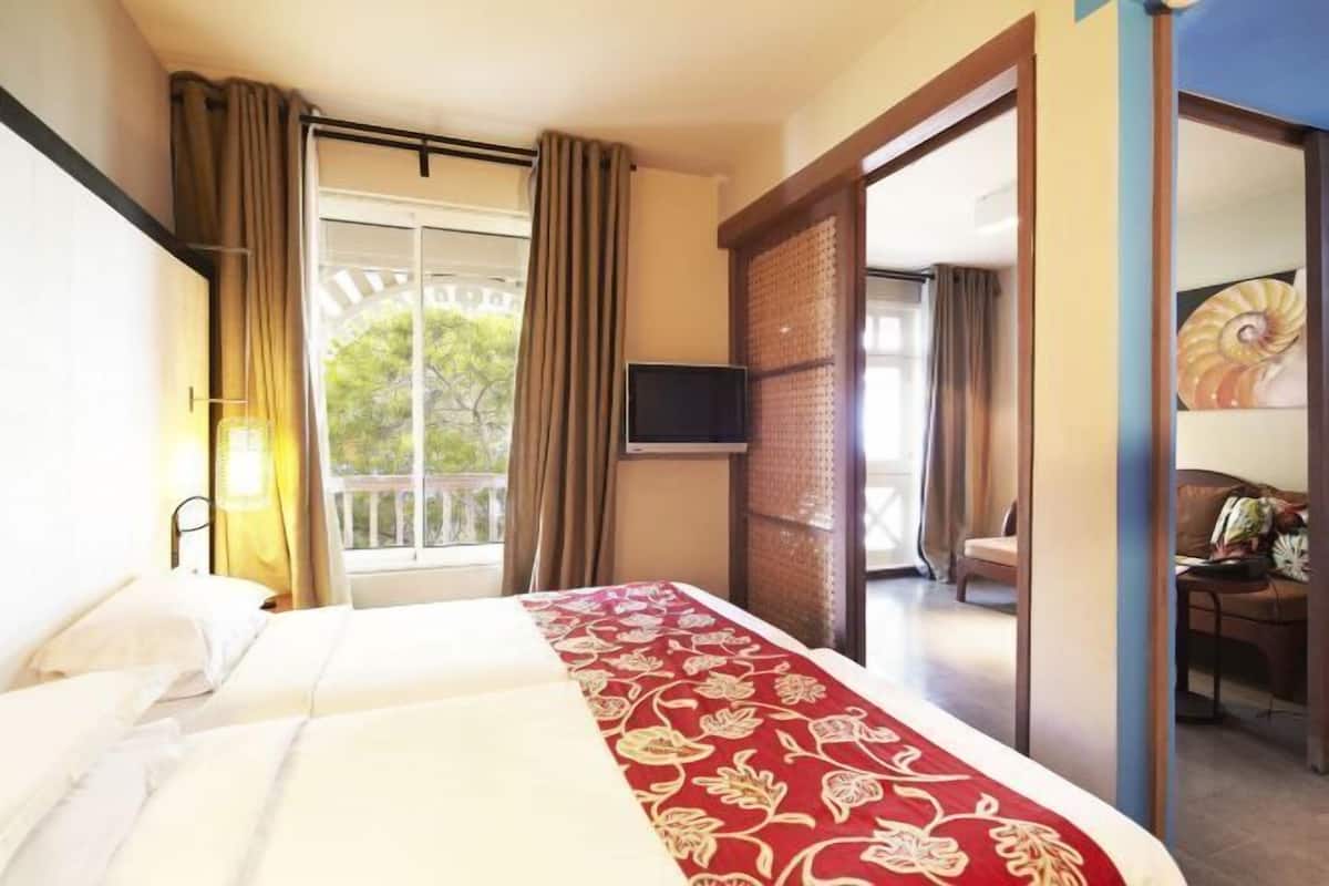 Habitación de hotel con cama, televisor, balcón con cortinas y vistas a zonas verdes.