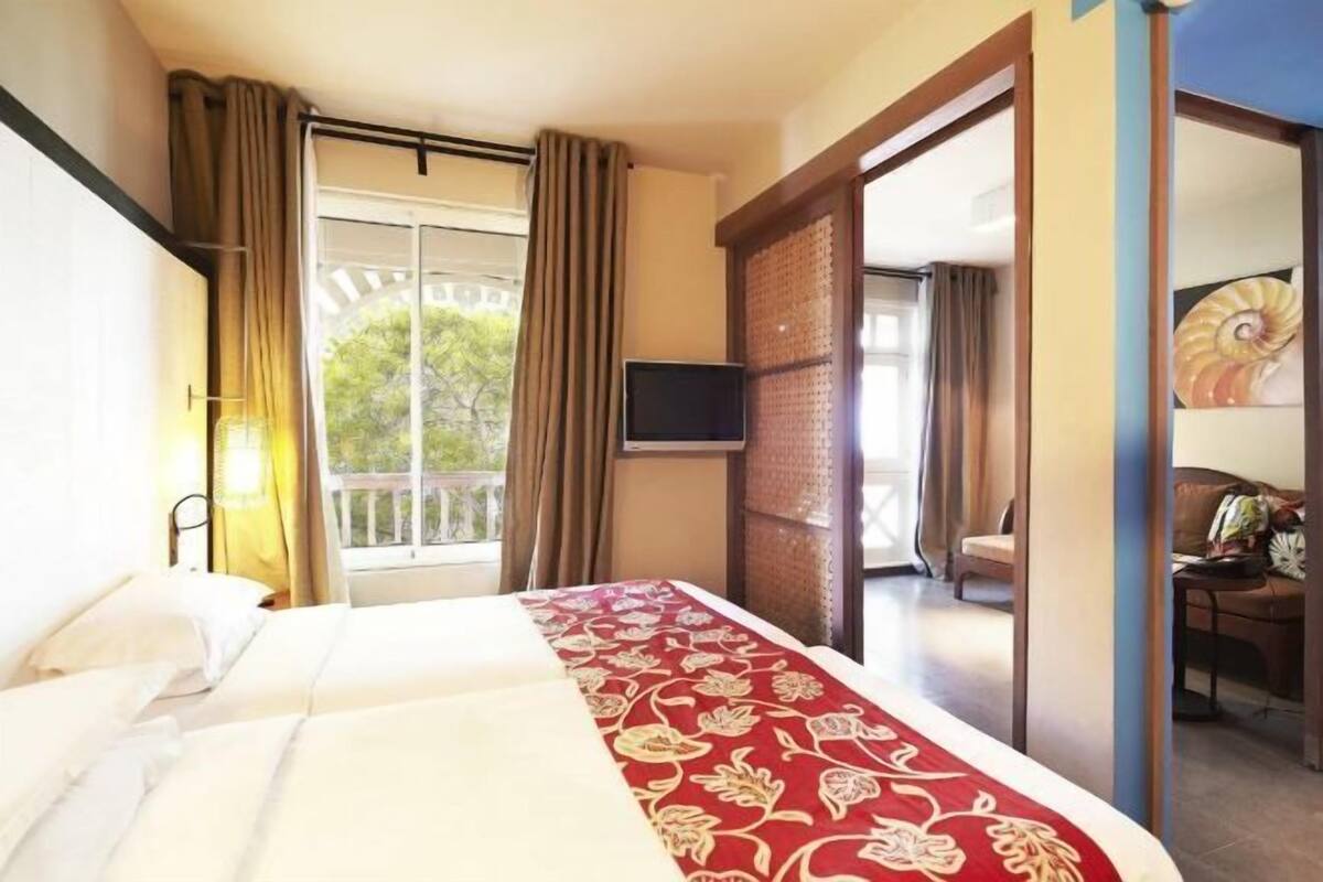 Habitación de hotel con cama, televisor, balcón y vistas a zonas verdes.