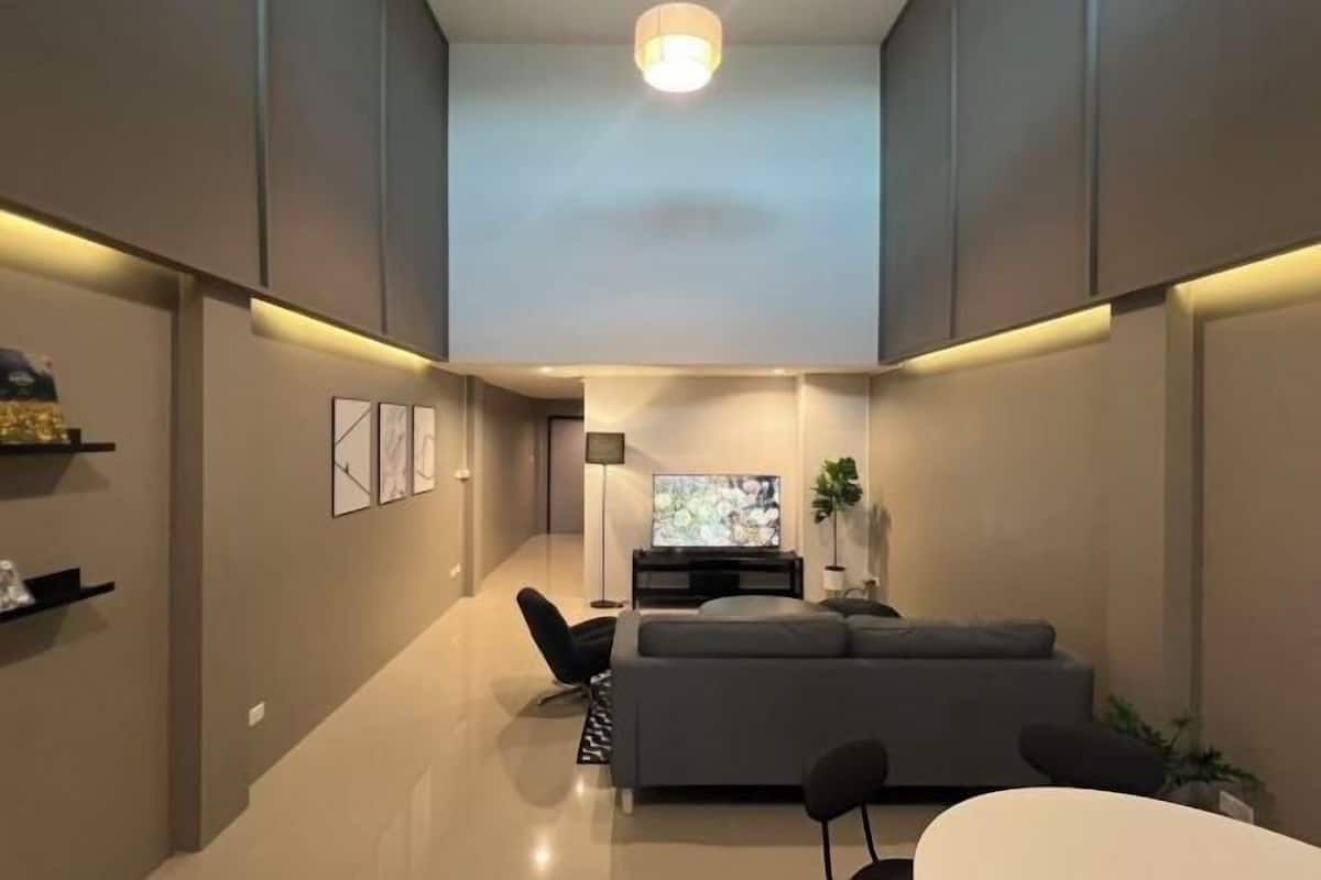 A modern living room with a grey sofa, black chairs, a flat-screen TV, and a ceiling light.