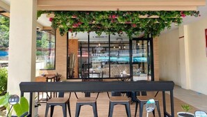 A modern indoor dining area with a long table and several stools, a brick wall, and a glass partition with a view of the outside. - Mildmint Self Serviced (Phuket)
