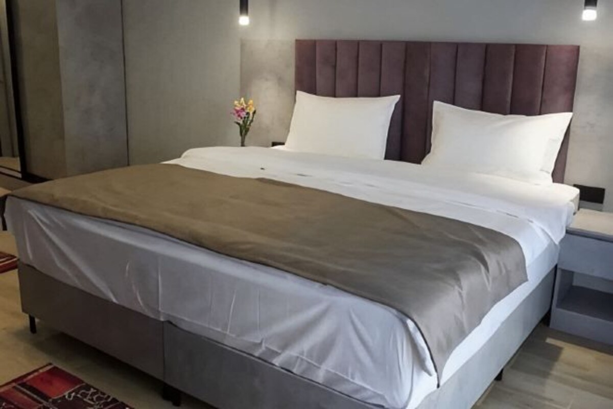 Un letto rifatto con cura, una panca da notte con un vaso di fiori e un comodino con una lampada.