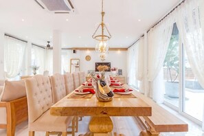A dining area with a long wooden table, cushioned chairs, and a chandelier. - 9 bedroom Stylish 2-Villa Combination by Hi At Home (Pattaya)