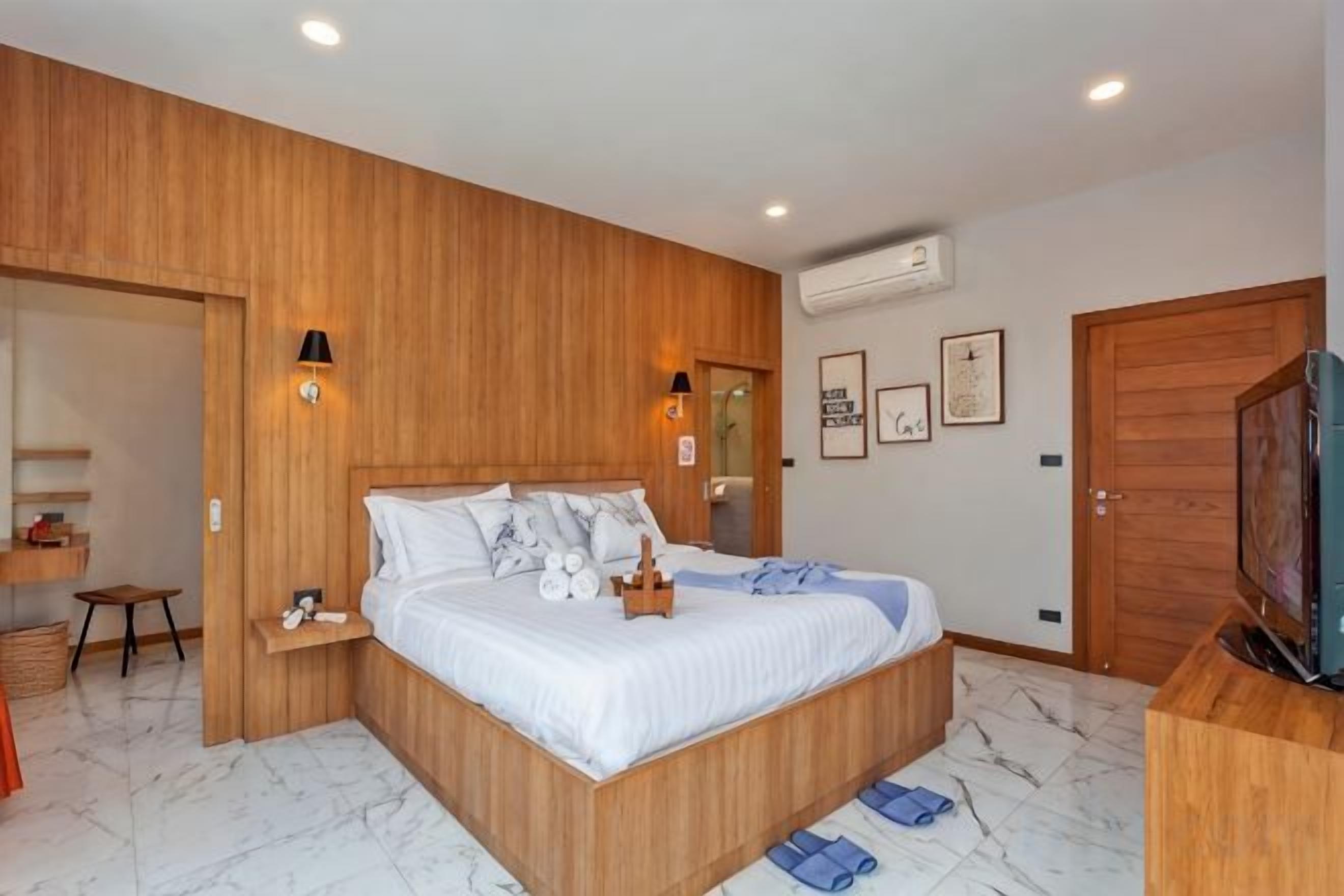 A bedroom with a wooden headboard, a bed with white bedding, a small bedside table, a television, and a wooden wardrobe.