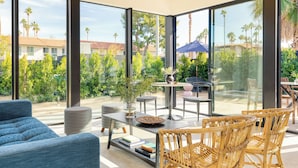 A modern living room with large glass windows, a blue sofa, wicker chairs, and a small table with a vase and books. - Sleeps 40 | Entire 16-Room Hotel, Pool | Blackhaus (Palm Springs)