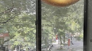A view through a window with horizontal blinds, a hanging light fixture, and a street scene with trees and buildings. - OH Trendy homes Hoan Kiem LTK Bancony City view (Hanoi)