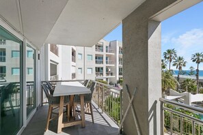 A balcony with a table and chairs, overlooking a building with multiple balconies and a view of palm trees and the ocean.
