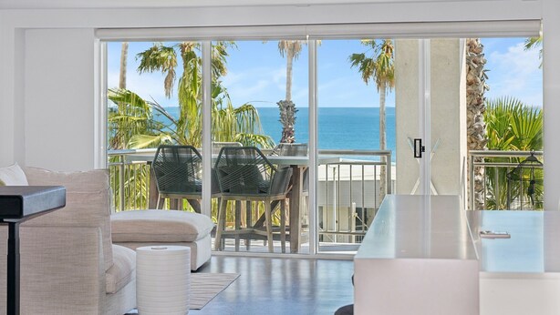 A modern living room with a large sliding glass door leading to a balcony with a view of the ocean and palm trees.