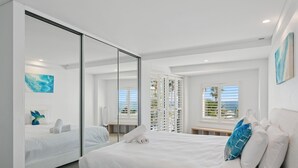 A modern hotel room with a large bed, a glass partition, a balcony with a view, and a painting on the wall.