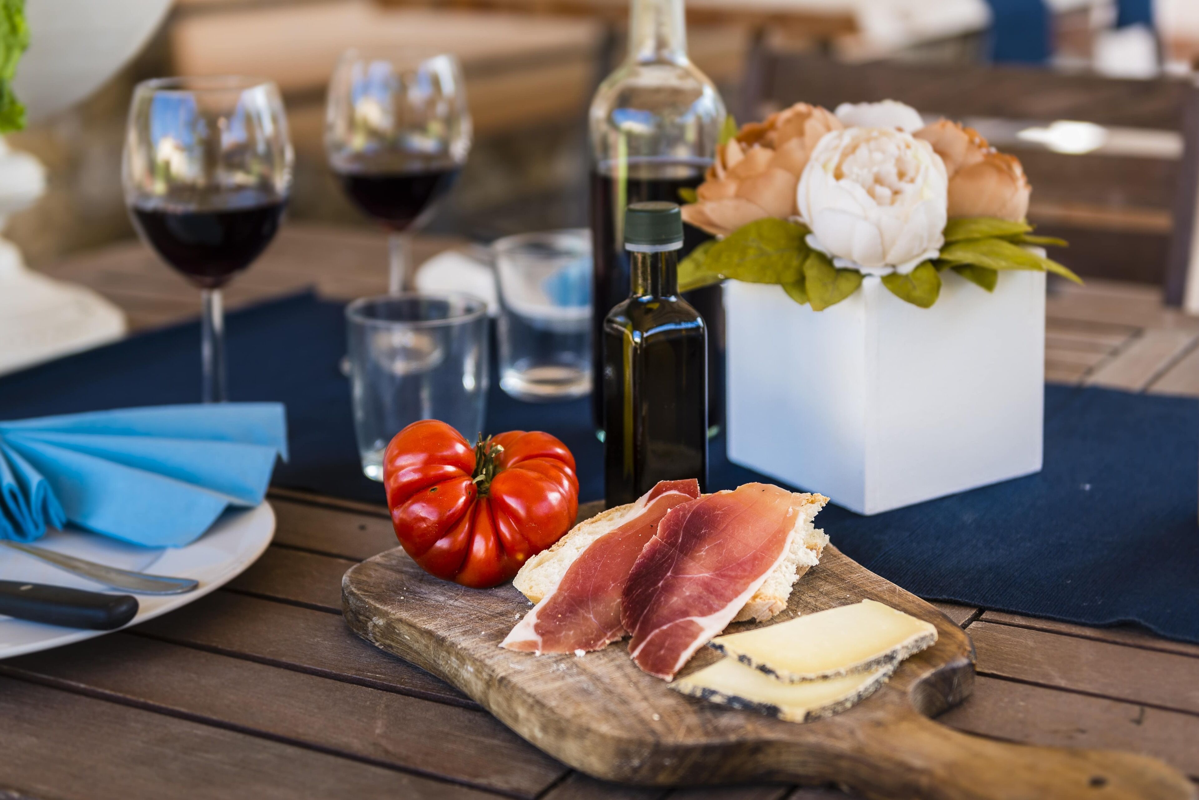 Una mesa de madera con una tabla de quesos, un tomate y un ramo de flores.