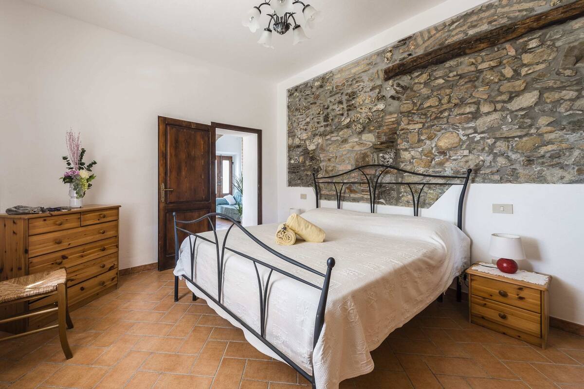 Een slaapkamer met een stenen muur, een bed met metalen frame, een houten dressoir, een stoel en een nachtkastje met een lamp.