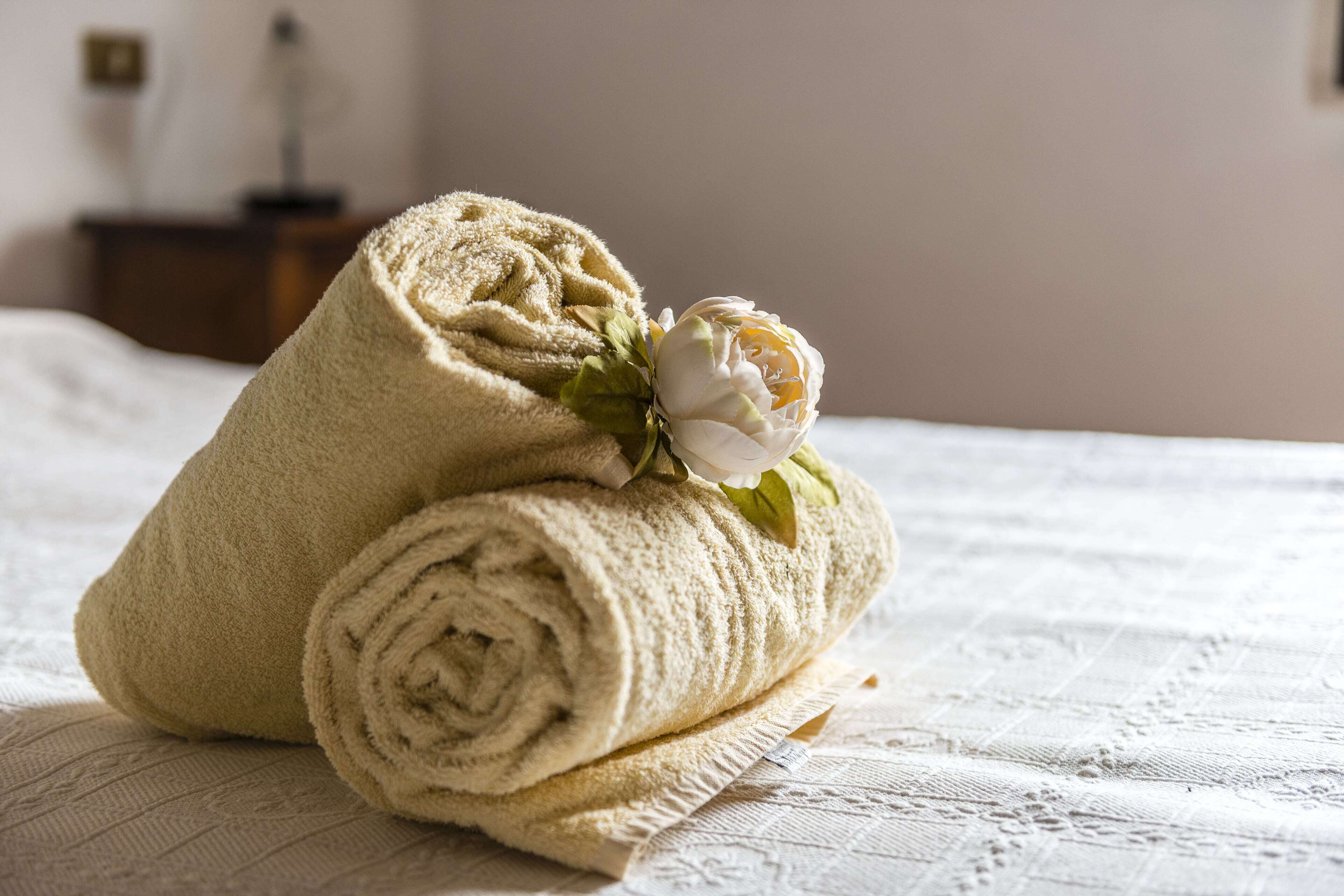 En prydligt vikt, beige handduk med en vit blomma ligger på en säng med ett mönstrat, vitt överkast.