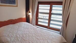 A bed with a white quilt, a wooden headboard, a nightstand with a lamp, and a window with curtains.