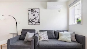 A modern living room with a grey sofa set, a floor lamp, a wall-mounted air conditioner, and a window with a view. - Arai 1chome (Tokyo)