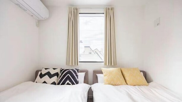 A room with two beds, a window with curtains, and an air conditioning unit on the wall. - Arai 1chome (Tokyo)