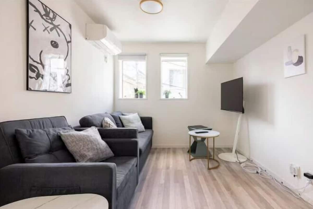 A modern living room with a grey sofa, a small round table, a flat-screen TV, and two windows with plants.