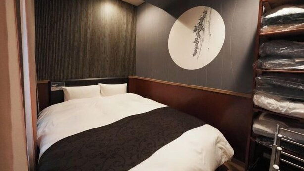 A hotel room with a bed, a wall-mounted circular artwork, and a shelving unit with folded linens. - Prosupa 5th #501 (Nagoya)
