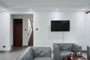 Ein modernes Wohnzimmer mit einem grauen Sofa, einem Couchtisch mit Dekoration, einem an der Wand befestigten Flachbildfernseher und einer Treppe, die in einen anderen Raum führt.