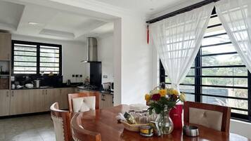 A dining area with a wooden table, chairs, a vase with flowers, and a view through large windows.