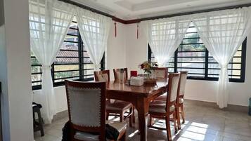 Un comedor con una mesa y sillas de madera, ventanas amplias con cortinas blancas y un suelo de baldosas.