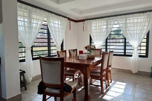 Uma sala de jantar com mesa e cadeiras de madeira, janelas amplas com cortinas brancas e piso ladrilhado.