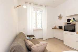 A compact living space with a sofa, a small kitchen area with an oven, and a window with curtains. - Colosseum Holidays - Marco Aurelio (Rome)