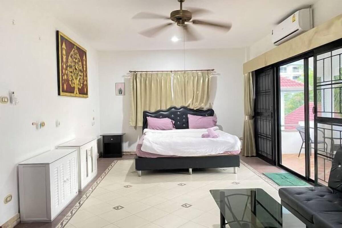 A bedroom with a bed, a ceiling fan, a radiator, a window with curtains, and a view of the outside.