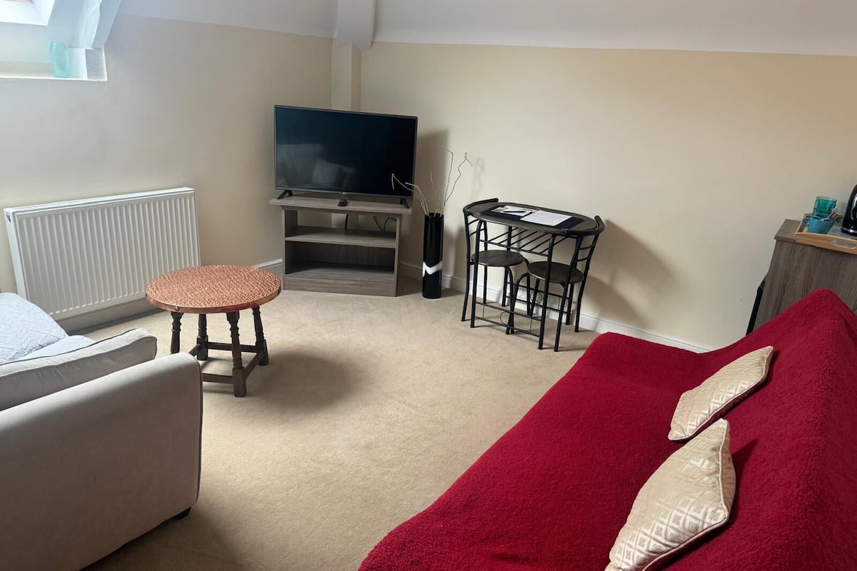 A living room with a sofa, a small round table, a TV on a stand, a radiator, and a window.