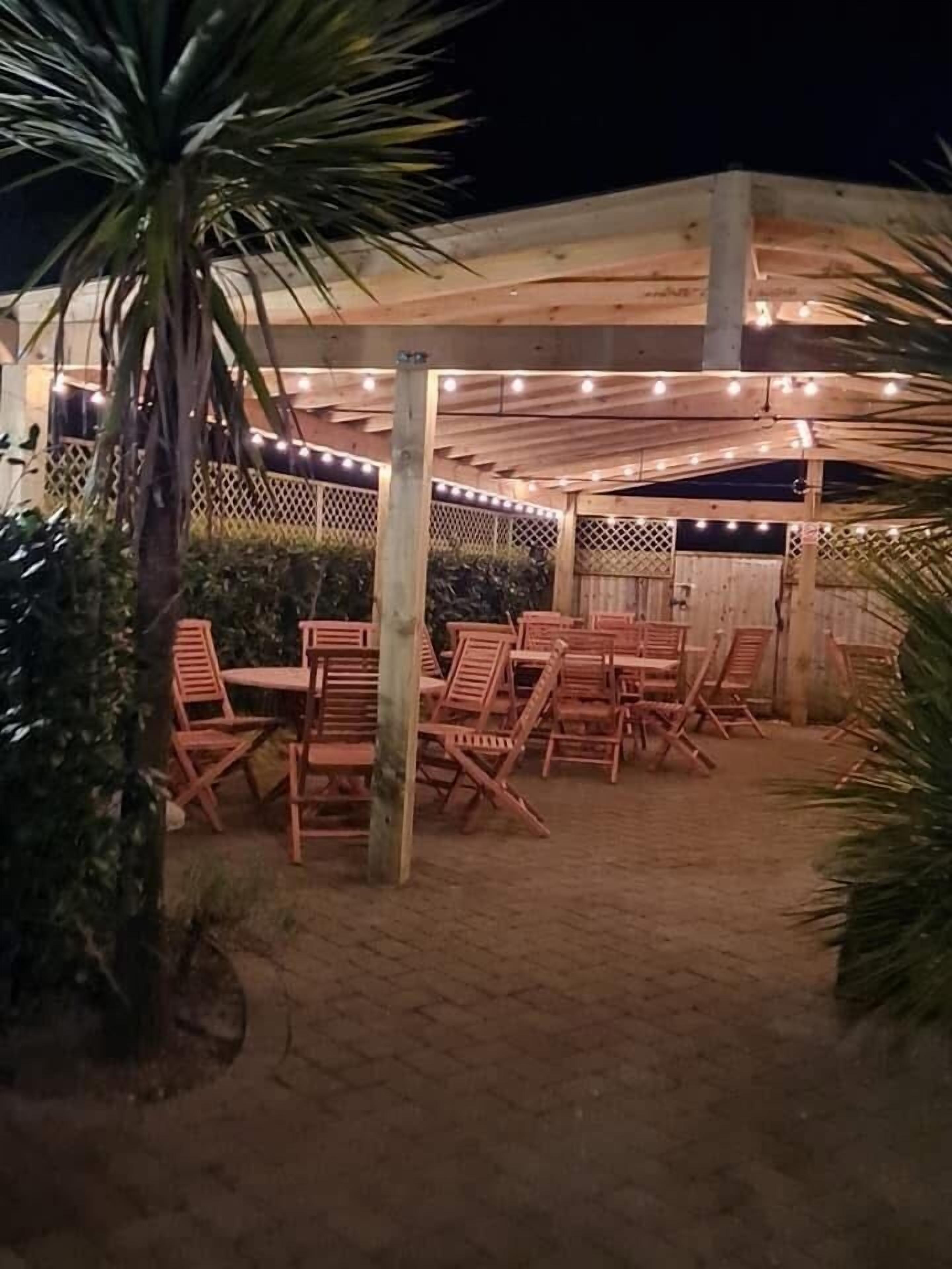 Un área de estar al aire libre con sillas y mesas de madera, rodeada por una pérgola de madera con luces colgantes.