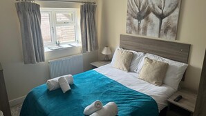 A hotel room with a bed, a nightstand, a lamp, a window with curtains, and a painting on the wall. - Greyhound Inn Freehouse  (Winterborne Kingston)