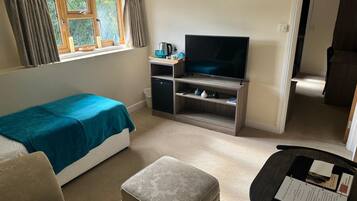 A small, well-lit room with a bed, a television on a wooden stand, a small cabinet, and a window with curtains.