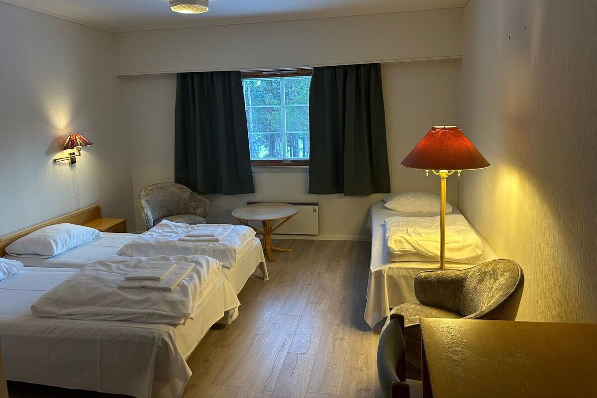 A hotel room with two single beds, a small round table, a chair, a lamp, and a window with curtains.