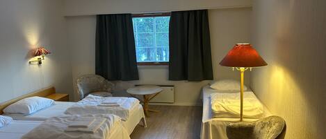 A hotel room with two single beds, a small round table, a chair, a lamp, and a window with curtains.