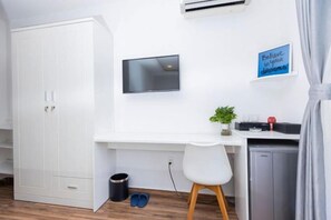 Une pièce avec une armoire blanche, un bureau avec une chaise, une petite plante en pot, un téléviseur et un panneau accroché au mur.