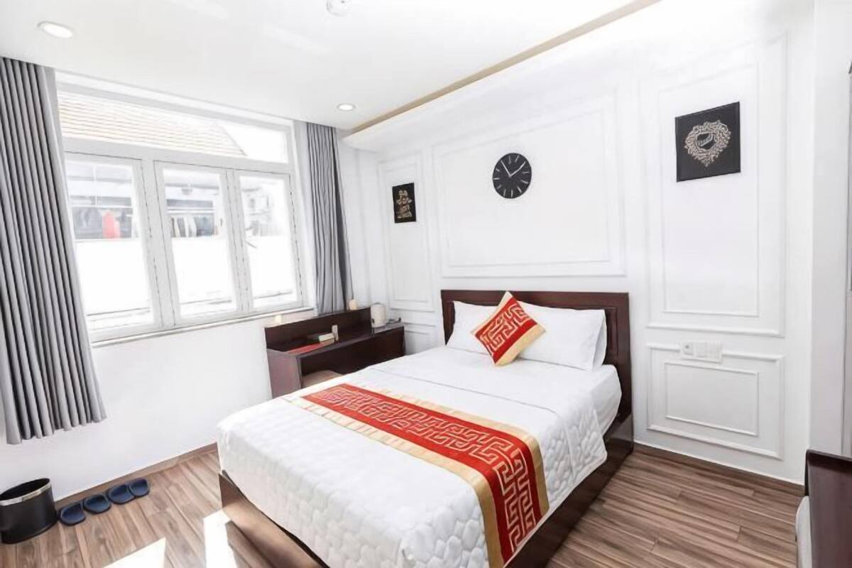 Ein Hotelzimmer mit einem großen Bett, einem Nachttisch, einer Uhr und einem Fenster mit Vorhängen.