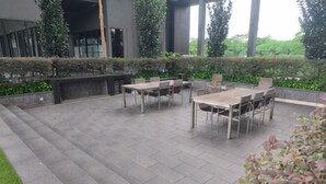 An outdoor patio area with a table and chairs, surrounded by greenery and a modern building in the background. - 8Pax/Green View next to KL118 ChinaTown & Train (Kuala Lumpur)