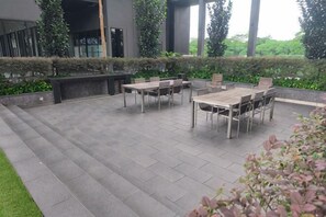 Un espace extérieur aménagé en terrasse, avec une table et des chaises, entouré de verdure et avec un bâtiment moderne en arrière-plan.