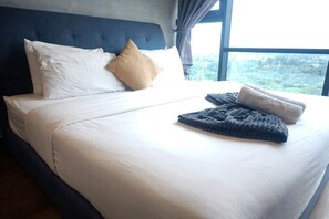 Una cama bien hecha con ropa blanca y una manta gris, un cabecero oscuro y una ventana con vistas a la vegetación.