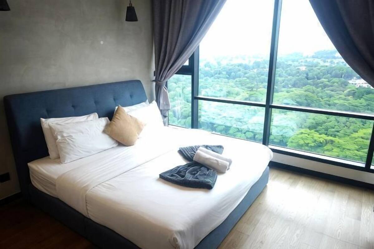 A bed with white linens, a blue headboard, and a view of greenery through large windows.