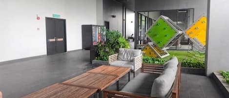 A modern lounge area with a sofa, a wooden table, and a small seating area. There are plants and a large window in the background.