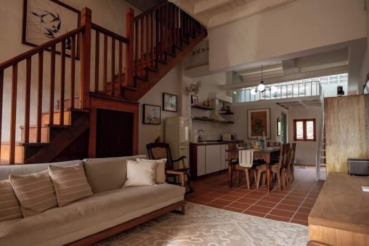 A living room with a sofa, wooden staircase, kitchen, dining area, and a view of the outside.